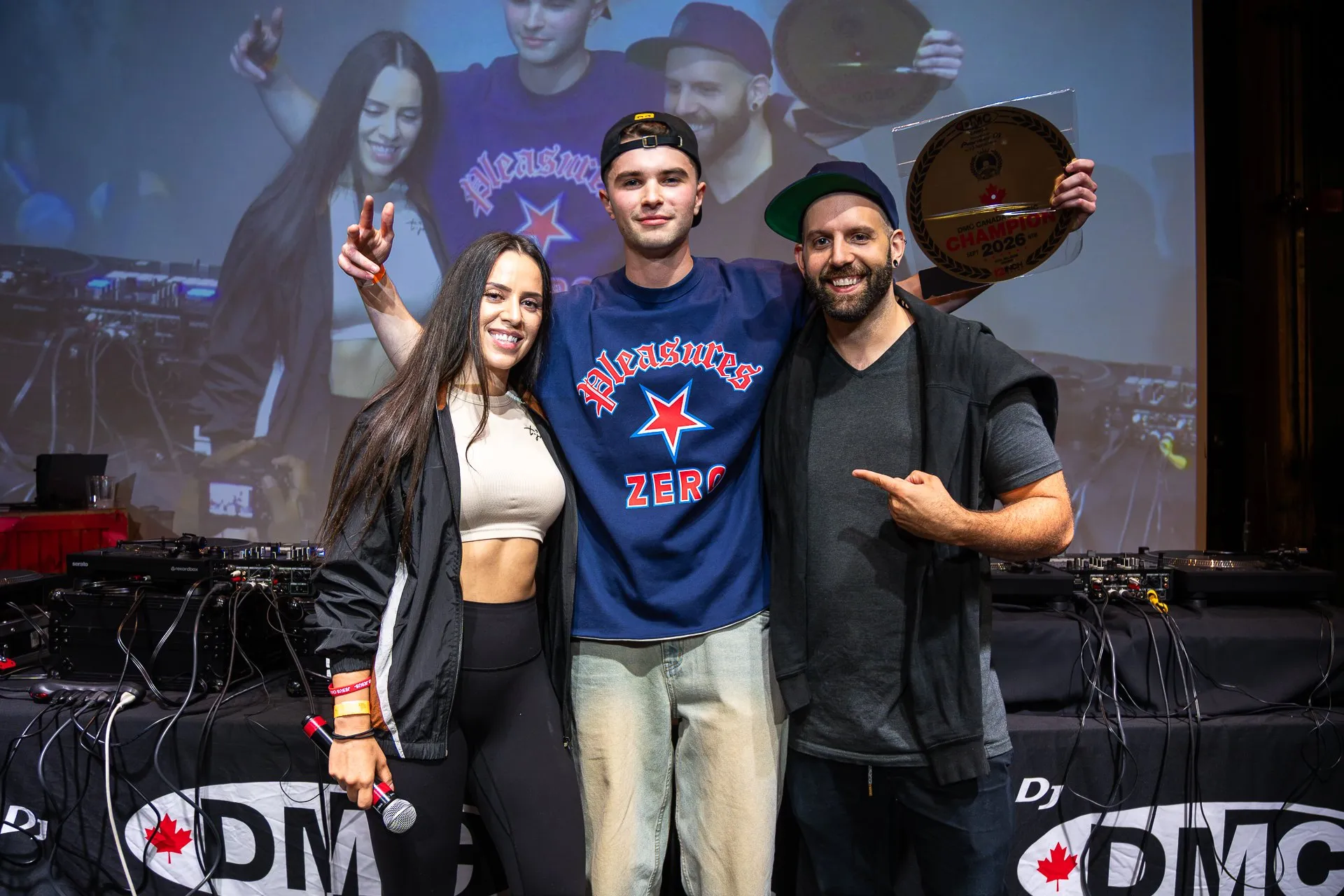 DEEJAY T-JR. on stage with the 2026 DMC Canadian champion, who holds up the winner’s plaque, in front of the DMC Canada table.