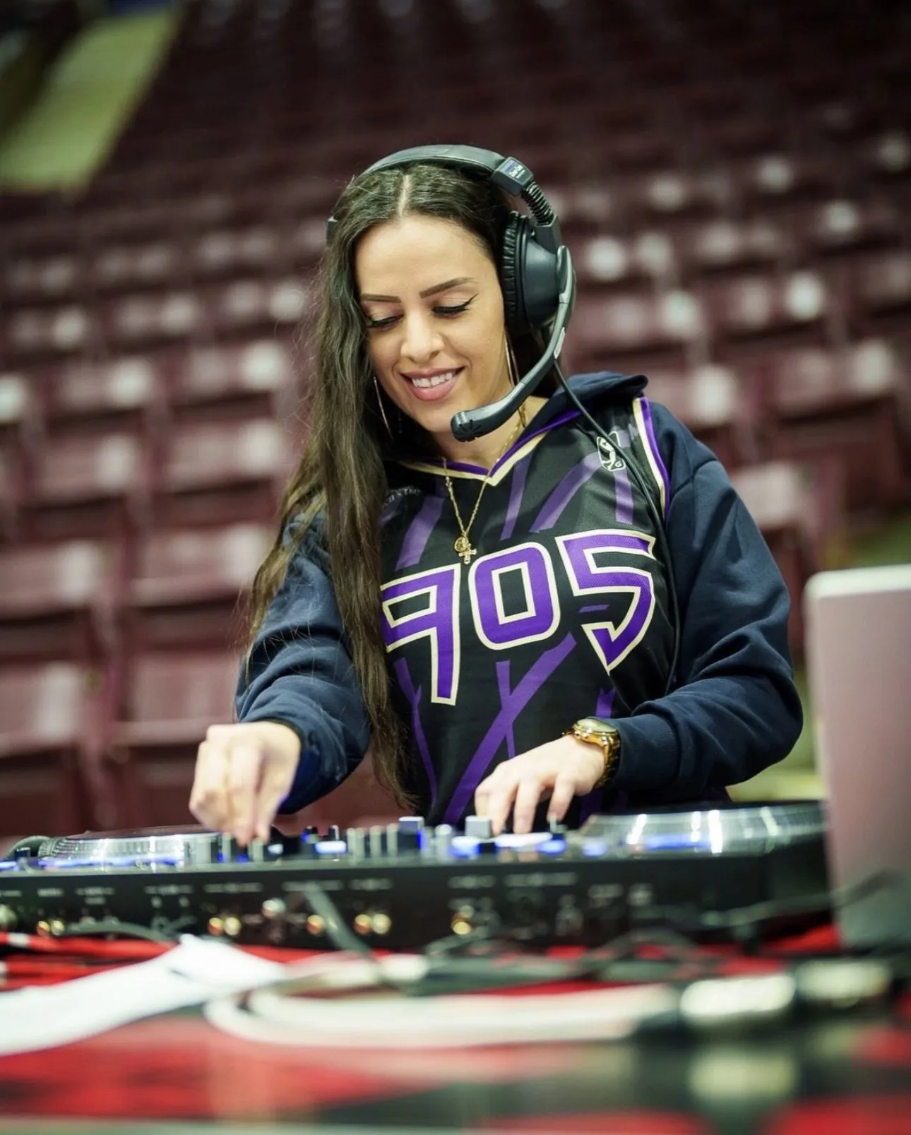 DEEJAY T-JR. on the decks courtside in a Raptors 905 jersey, wearing a headset, empty arena seats behind her.